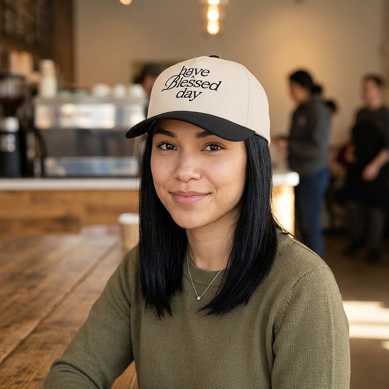 A woman with dark hair wears a cream and black baseball cap embroidered with "have a Blessed day" while sitting at a wooden table in a cafe.