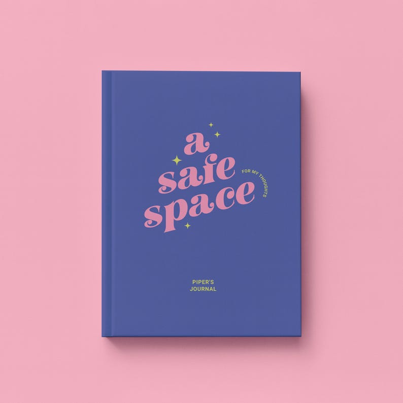 May include: A hardcover journal with a blue cover and the words "a safe space" in pink script. The text "for my thoughts" is in a smaller font, and the name "Piper's Journal" is at the bottom. The background is pink.