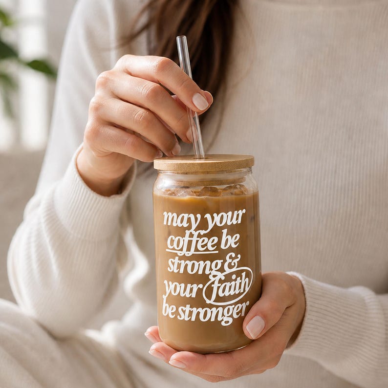 May include: A clear glass coffee cup filled with coffee and ice, featuring a bamboo lid and a clear straw. The cup has white text that reads "may your coffee be strong & your faith be stronger".