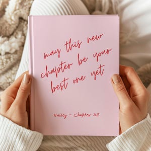 Puede incluir: Un libro de tapa dura de color rosa claro con texto rojo en cursiva que dice "may this new chapter be your best one yet" y "Hailey - Chapter 30" impreso en una fuente más pequeña en la parte inferior. El libro tiene un acabado mate.