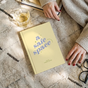May include: A light yellow journal with the words "a safe space" in blue script. The journal also has the text "for my thoughts" and "Lola's Therapy Notes". A glass of liquid sits nearby on a patterned rug.