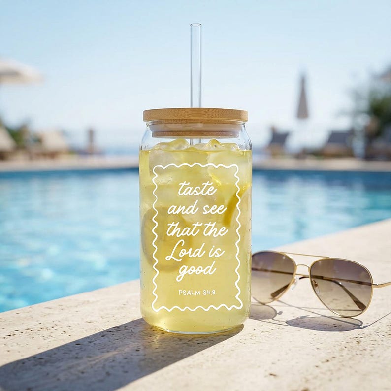 May include: A clear glass tumbler with a bamboo lid and a glass straw, filled with a yellow beverage and ice. The tumbler has white text that reads "taste and see that the Lord is good" and is next to a pair of gold-rimmed sunglasses.