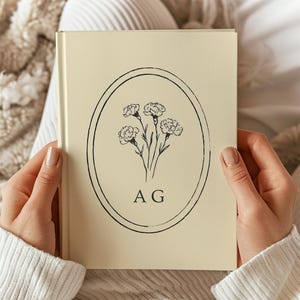 Op de afbeelding: Een crèmekleurig hardcover boek met een zwarte lijntekening van een klein boeket anjers, gecentreerd in een dun ovaal kader. Onder het bloemmotief staan de hoofdletters A en G gedrukt in een eenvoudig zwart schreeflettertype.