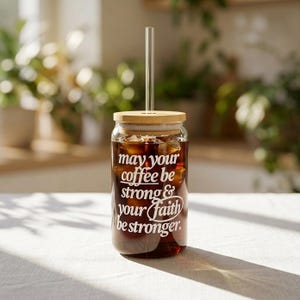 May include: A clear glass tumbler filled with iced coffee, featuring a bamboo lid and a glass straw. The tumbler has white text that reads "may your coffee be strong & your faith be stronger."