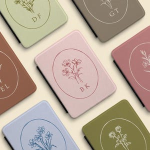 May include: A collection of rectangular e-reader cases in various muted colors including sage green, dusty rose, terracotta, and slate blue. Each case features a centered oval outline containing a line-art floral illustration and two-letter initials.
