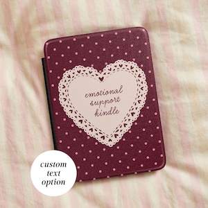May include: A dark maroon e-reader case featuring a white polka dot pattern and a central cream-colored heart-shaped doily graphic. The text "emotional support kindle" is printed in dark maroon script inside the heart. A white circular label reads "custom text option".