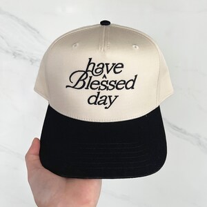 A hand holds a cream and black baseball cap embroidered with "have a Blessed day" against a light marble background.