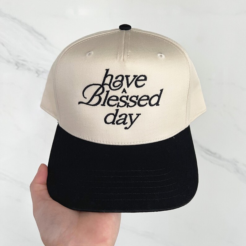 A hand holds a cream and black baseball cap embroidered with "have a Blessed day" against a light marble background.