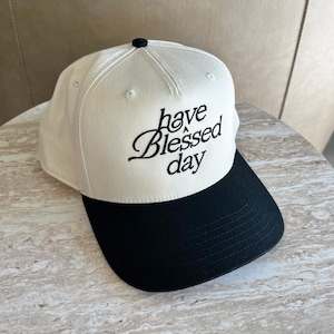 A two-tone baseball cap with a black brim and a cream crown, embroidered with "have a Blessed day" in black script, resting on a light-colored surface.