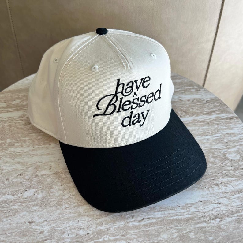 A two-tone baseball cap with a black brim and a cream crown, embroidered with "have a Blessed day" in black script, resting on a light-colored surface.