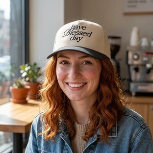 A smiling woman with red hair wears a cream and black baseball cap embroidered with "have a Blessed day" in a cafe.