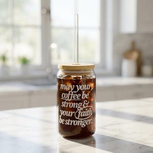 May include: A clear glass coffee cup filled with iced coffee, a glass straw, and a wooden lid. The cup has white text that reads "may your coffee be strong & your faith be stronger."