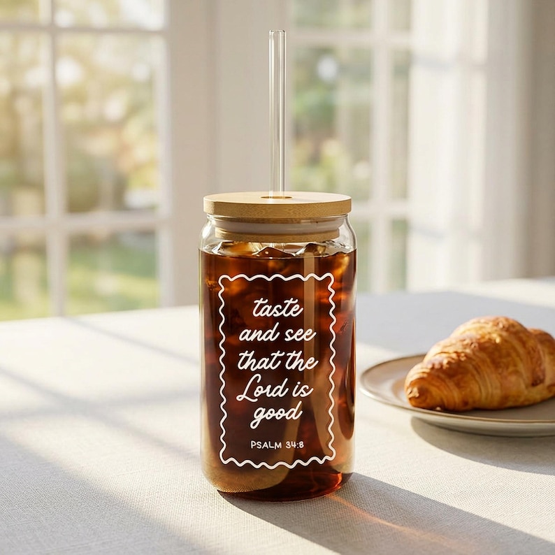 May include: A clear glass tumbler filled with a dark liquid, featuring a wooden lid and a clear straw. The tumbler has white text that reads "taste and see that the Lord is good" with "Psalm 34:8" below. A croissant sits on a plate.