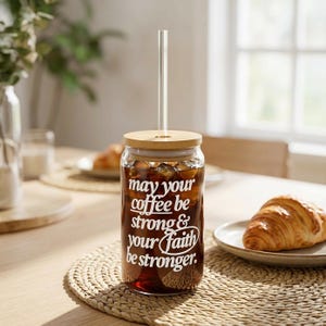 May include: A clear glass tumbler filled with iced coffee, featuring a bamboo lid and a clear straw. The tumbler has white text that reads "May your coffee be strong & your faith be stronger." The tumbler sits on a woven placemat.