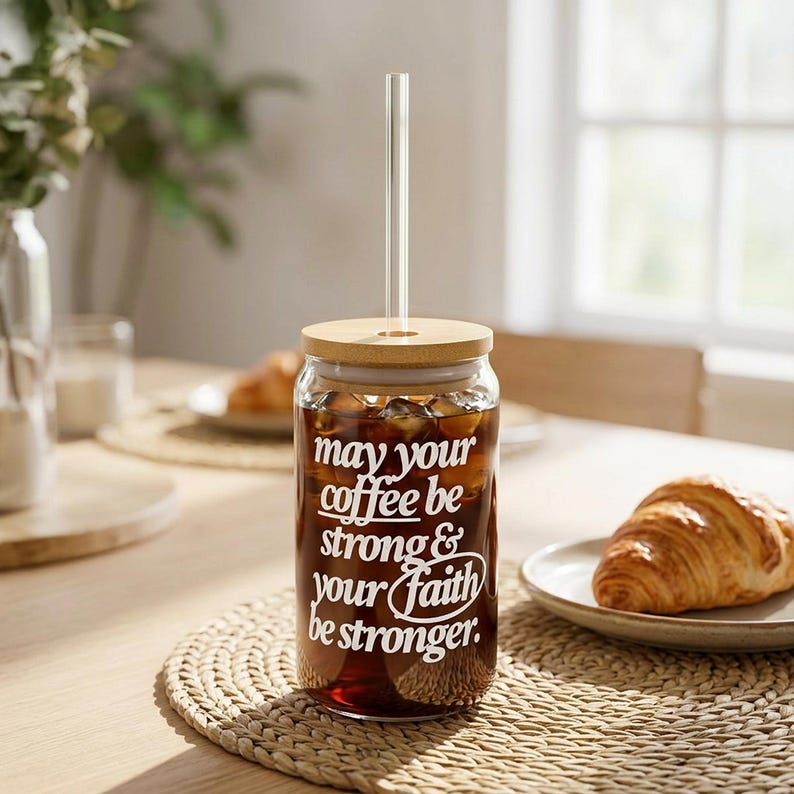 May include: A clear glass tumbler filled with iced coffee, featuring a bamboo lid and a clear straw. The tumbler has white text that reads "May your coffee be strong & your faith be stronger." The tumbler sits on a woven placemat.