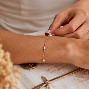 May include: A delicate gold chain bracelet with small white pearl accents. The bracelet is shown on a person's wrist.
