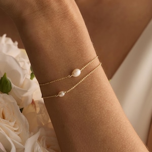 Peut inclure: Deux bracelets fins en or, chacun orné d'une petite perle, sont portés au poignet d'une personne. Les bracelets sont superposés. Des fleurs blanches et une robe blanche sont en arrière-plan. Le texte "HandmadeByCozayh" est visible.