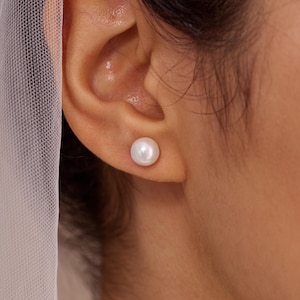 Può includere: Primo piano di un orecchio con un singolo orecchino a bottone di perla bianco e rotondo. L'orecchino è centrato sul lobo dell'orecchio. Lo sfondo è sfocato, con un tessuto bianco trasparente visibile a sinistra. Il testo "HandmadeByCozayh" è in basso a destra.