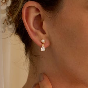 Pode incluir: Um close-up de um brinco dourado com um pequeno pino de pérola e uma pedra preciosa transparente em forma de gota. O brinco é usado em uma orelha, com um fundo desfocado. O texto "HandmadeByCozayh" é visível no canto inferior direito.