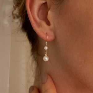 May include: Gold-tone drop earrings featuring two white pearls. The earrings are suspended from a delicate gold hook, with a smaller pearl above a larger one. The design is simple and elegant, suitable for special occasions.