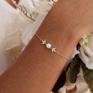 Peut inclure: Un bracelet en or fin orné d'une perle centrale et de deux motifs en forme de feuilles sertis de cristaux étincelants. Le bracelet est porté au poignet, avec un bouquet de fleurs blanches en arrière-plan.