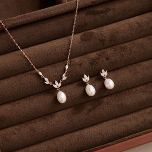 May include: Rose gold necklace and earrings set featuring a pear-shaped pearl pendant and matching earrings. The necklace has a delicate chain with leaf-shaped accents. The earrings have a pearl drop design. The jewelry is displayed on a brown velvet surface.