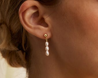 May include: Gold-colored drop earrings featuring three white pearls. The earrings are suspended from a small gold ball and are designed to dangle. The jewelry is shown on a person's ear, highlighting the elegant design.