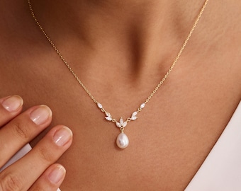 May include: A delicate gold necklace featuring a pear-shaped pearl pendant. The necklace has a fine chain and is adorned with small, sparkling accents. The design includes leaf-like details, creating an elegant and refined aesthetic. The necklace is displayed on a person.