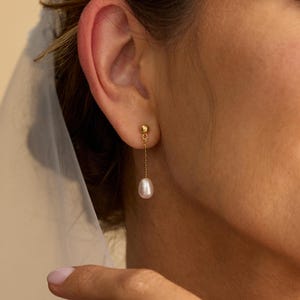 Op de afbeelding: Een gouden oorbel met een kleine bolvormige stud en een fijne ketting die een witte, druppelvormige parel vasthoudt. De oorbel wordt op een oor gedragen, tegen een zachte achtergrond.