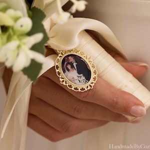 May include: An oval gold-toned metal photo charm featuring a decorative filigree border, attached to a cream-colored satin ribbon wrapped around a bouquet handle. The charm displays a small photograph of a couple in formal wedding attire.