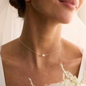 May include: A delicate gold necklace featuring a single, lustrous pearl pendant. The necklace is worn around the neck, with a focus on the pearl. The background is soft and blurred, highlighting the jewelry. The necklace is a simple, elegant piece.