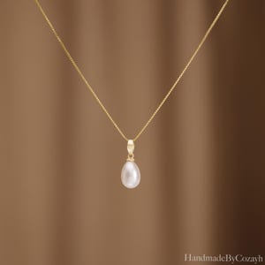 May include: A delicate gold box chain necklace featuring a single teardrop-shaped white pearl pendant suspended from a small gold bail. The necklace is centred in the frame, showcasing the smooth lustre of the pearl and the fine link chain design.