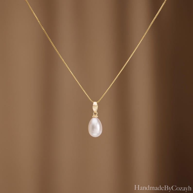 May include: A delicate gold box chain necklace featuring a single teardrop-shaped white pearl pendant suspended from a small gold bail. The necklace is centred in the frame, showcasing the smooth lustre of the pearl and the fine link chain design.