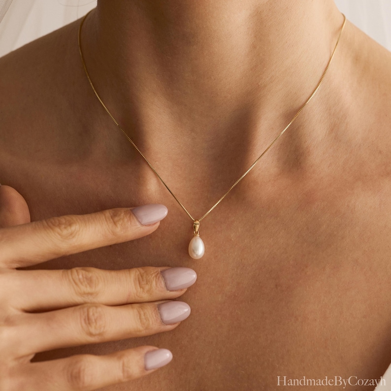 May include: A delicate gold chain necklace featuring a single, teardrop-shaped white pearl pendant. The pendant is suspended from a simple gold bail, and the chain rests against the collarbone, showcasing a classic and refined jewellery design.