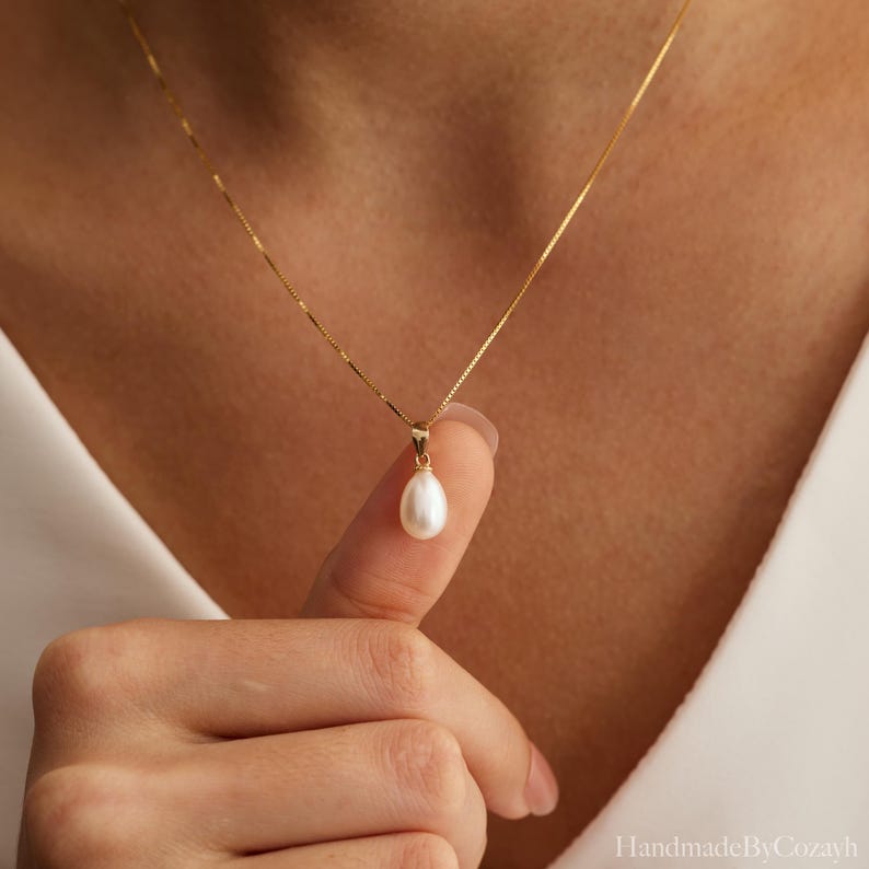 May include: A delicate gold chain necklace featuring a single, smooth, white teardrop-shaped pearl pendant. The pendant is suspended from a thin, polished gold bail, creating a refined and classic piece of jewellery that rests against the skin.