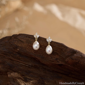 May include: A pair of gold-colored drop earrings featuring a pear-shaped white pearl suspended from a small, leaf-shaped cluster of clear crystals. The earrings are displayed on a dark brown wooden surface.