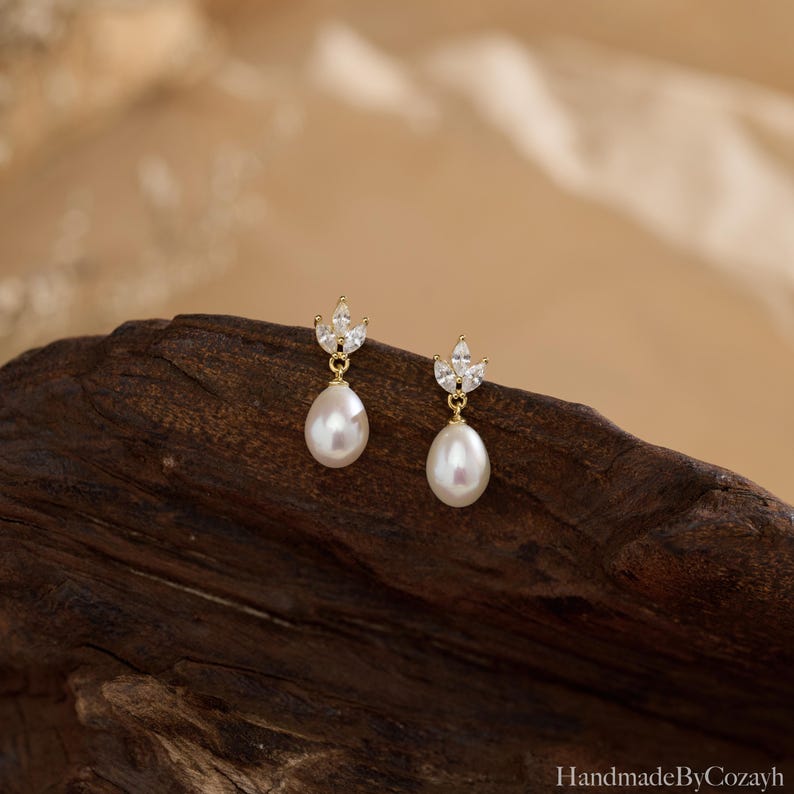 May include: A pair of gold-colored drop earrings featuring a pear-shaped white pearl suspended from a small, leaf-shaped cluster of clear crystals. The earrings are displayed on a dark brown wooden surface.