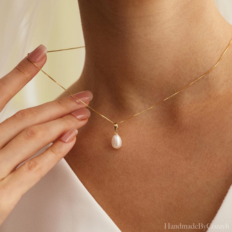 May include: A delicate gold chain necklace featuring a single teardrop-shaped white pearl pendant. The necklace is secured with a simple gold bail connecting the pearl to the thin, polished gold box chain that rests against the collarbone area.
