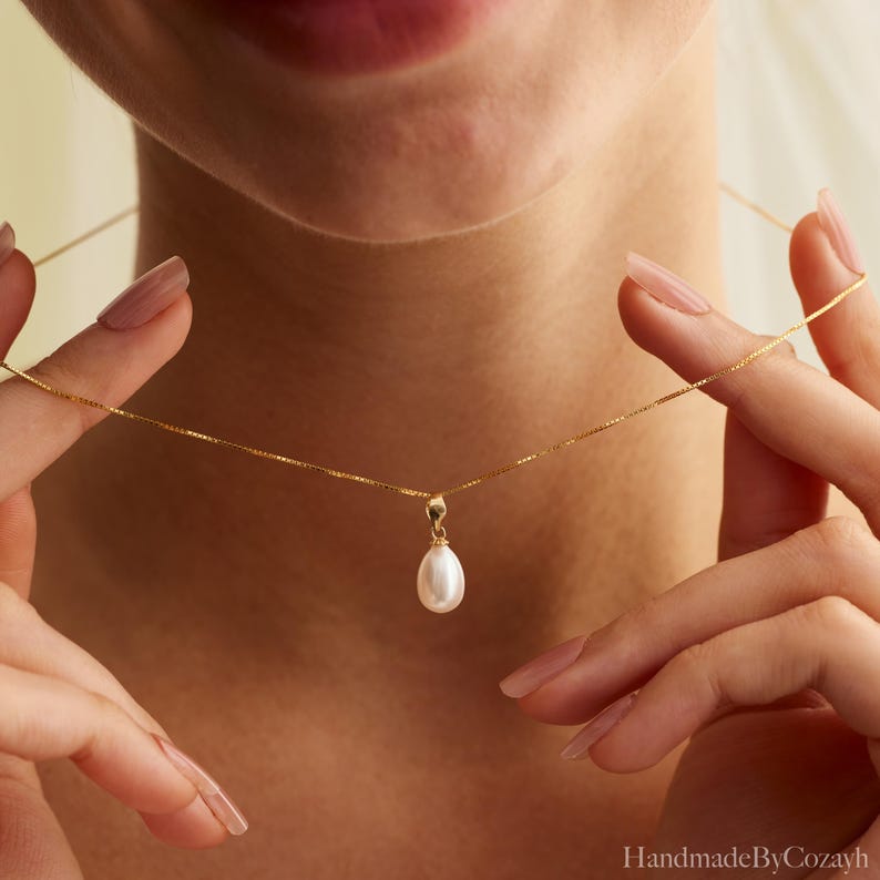 May include: A delicate gold chain necklace featuring a single, smooth, white teardrop-shaped pearl pendant. The pendant is suspended from a thin, polished gold link chain that rests against the neck, showcasing a simple and refined jewellery design.