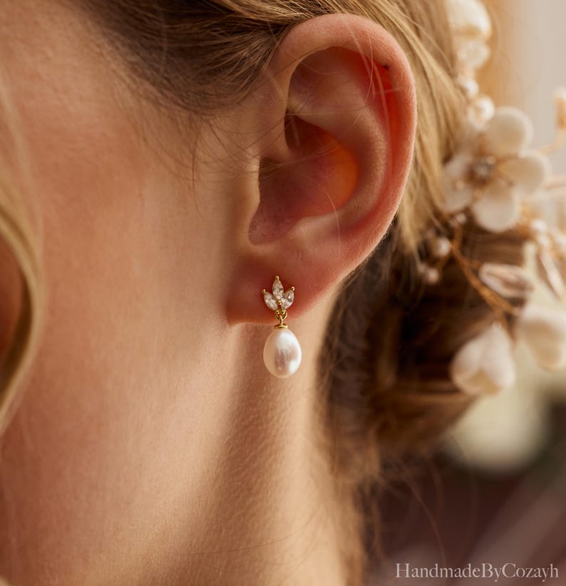 May include: A gold-toned earring with a teardrop pearl and a cluster of clear crystals. The earring is attached to the earlobe. The earring has a leaf-like design at the top. The earring is a piece of jewelry.