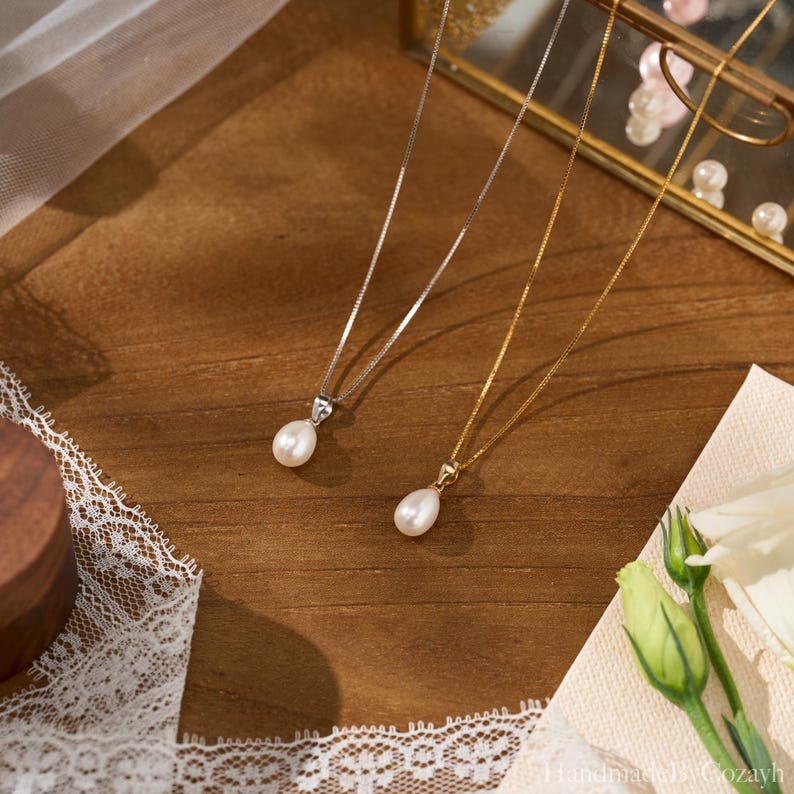 May include: Two delicate necklaces featuring teardrop-shaped white pearls suspended on thin metal chains. One necklace has a silver-toned chain and setting, while the other features a gold-toned chain and setting, both with a polished finish.