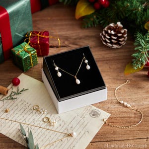 May include: A gold-toned jewelry set with white pearl accents, including a necklace and earrings, presented in a black and white box. A gold bracelet with pearls and other jewelry pieces are also visible on a wooden surface.