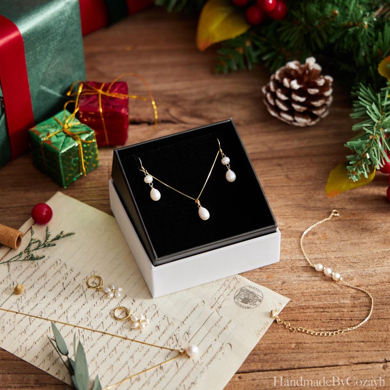 May include: A gold-toned jewelry set with white pearl accents, including a necklace and earrings, presented in a black and white box. A gold bracelet with pearls and other jewelry pieces are also visible on a wooden surface.