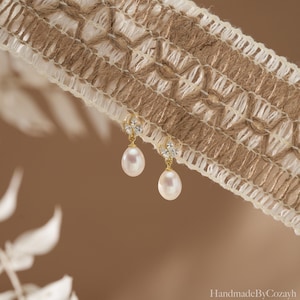 May include: A pair of gold-colored earrings with teardrop-shaped pearls. The earrings have a small, decorative cluster of clear stones at the top. The earrings are hanging in front of a brown and white woven fabric.
