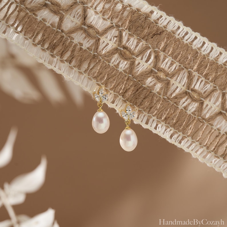 May include: A pair of gold-colored earrings with teardrop-shaped pearls. The earrings have a small, decorative cluster of clear stones at the top. The earrings are hanging in front of a brown and white woven fabric.