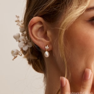 May include: Gold-toned drop earrings featuring a pear-shaped pearl suspended from a cluster of small, clear crystals. The earrings are worn on an ear, with a floral hair accessory visible in the background. The earrings are delicate and elegant.