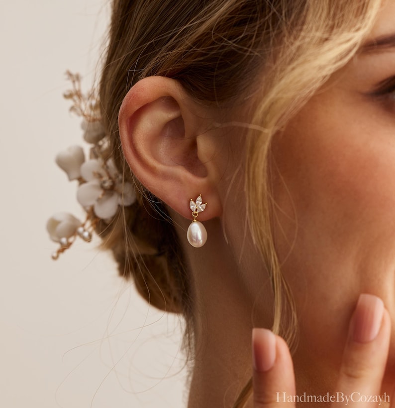 May include: Gold-toned drop earrings featuring a pear-shaped pearl suspended from a cluster of small, clear crystals. The earrings are worn on an ear, with a floral hair accessory visible in the background. The earrings are delicate and elegant.