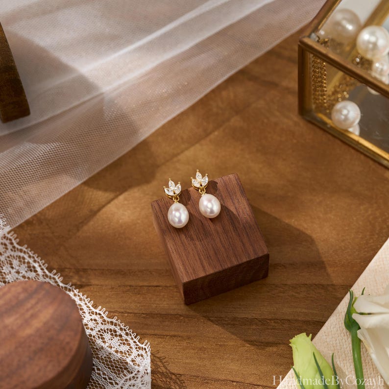 May include: A pair of gold-colored drop earrings with a pearl-shaped pendant. Each earring features a small, leaf-shaped design with clear, sparkling accents. The earrings are displayed on a small, dark wooden block.