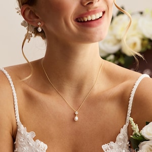 May include: A gold necklace with a small pendant featuring a pearl and a delicate leaf design. Matching gold earrings with pearls are also visible. The jewelry is worn by someone wearing a white lace dress, suggesting a bridal theme.