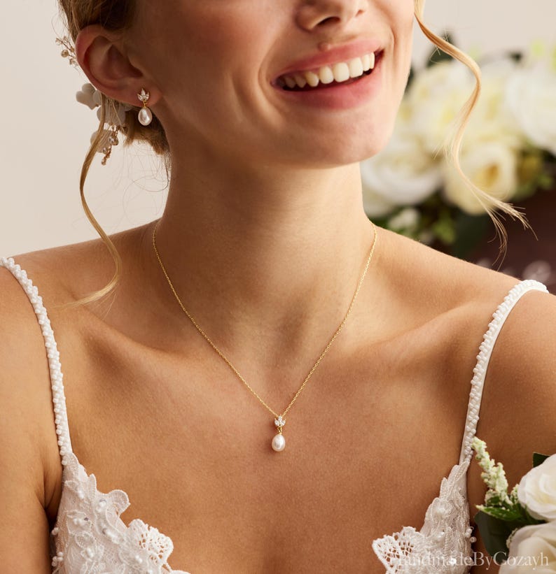 May include: A gold necklace with a small pendant featuring a pearl and a delicate leaf design. Matching gold earrings with pearls are also visible. The jewelry is worn by someone wearing a white lace dress, suggesting a bridal theme.